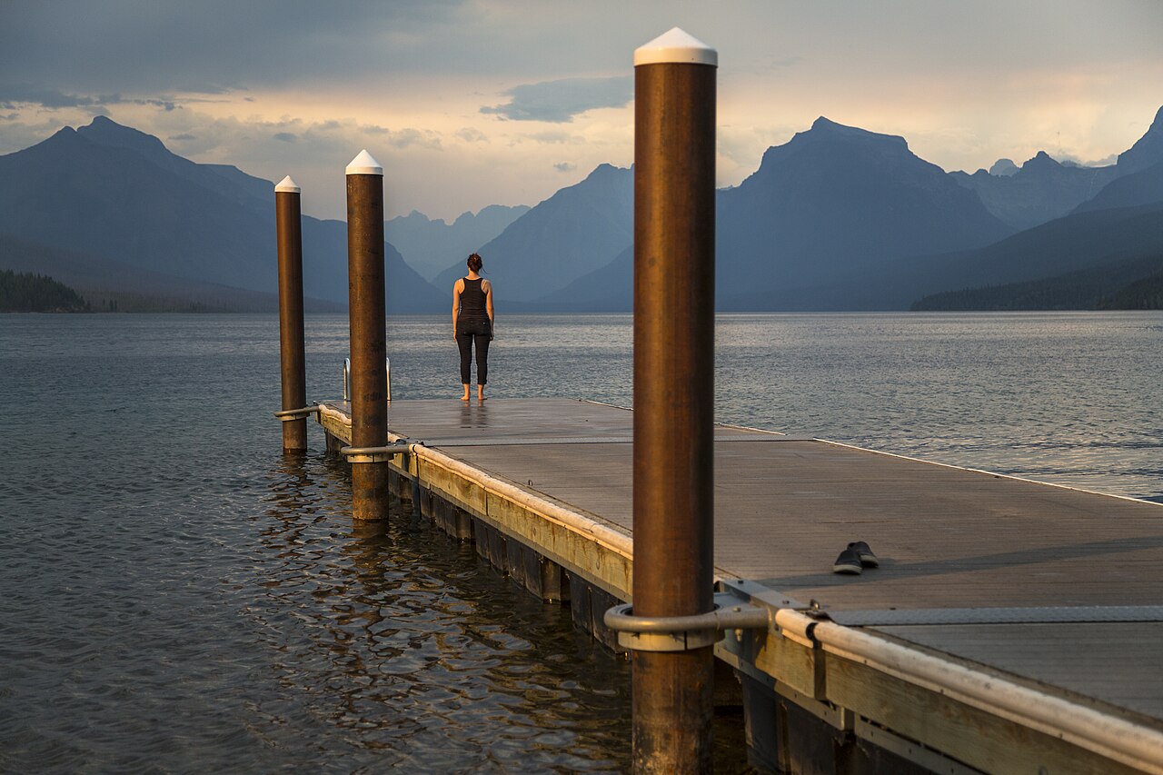 A person standing at the end of a dock overlooking Lake McDonald and the mountains